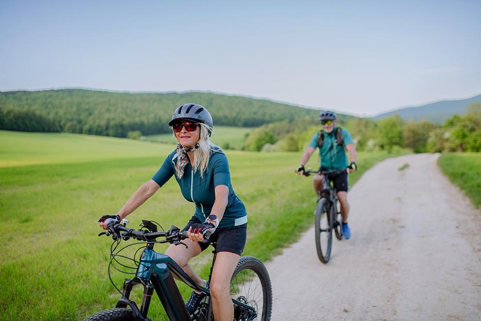 An active senior couple riding electric bicycles on path at summer park, healthy lifestyle concept.